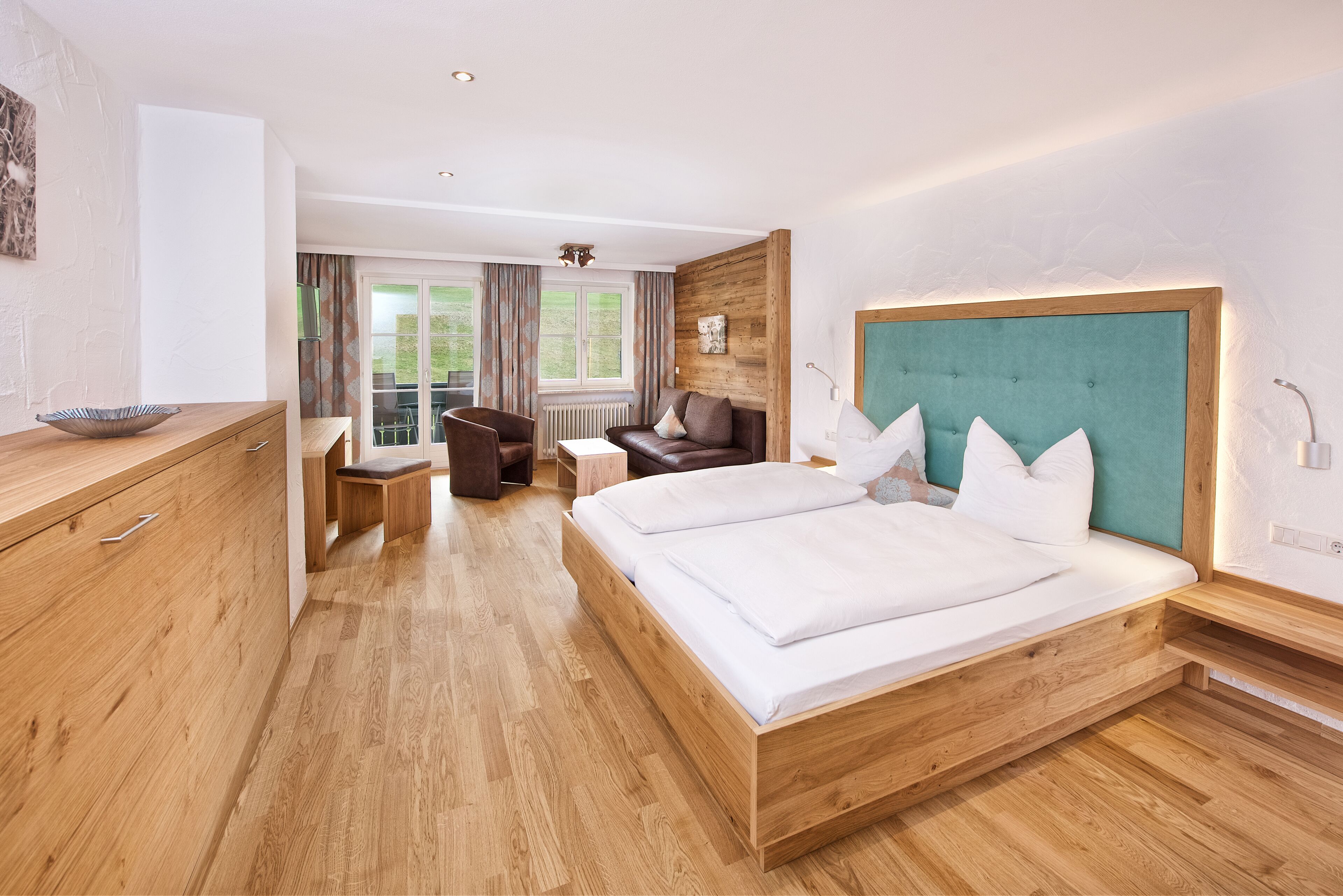 Una habitación de hotel moderna con una cama y cómoda de madera, un pequeño rincón de descanso con una silla y mesa, y vistas al exterior a través de un ventanal grande.