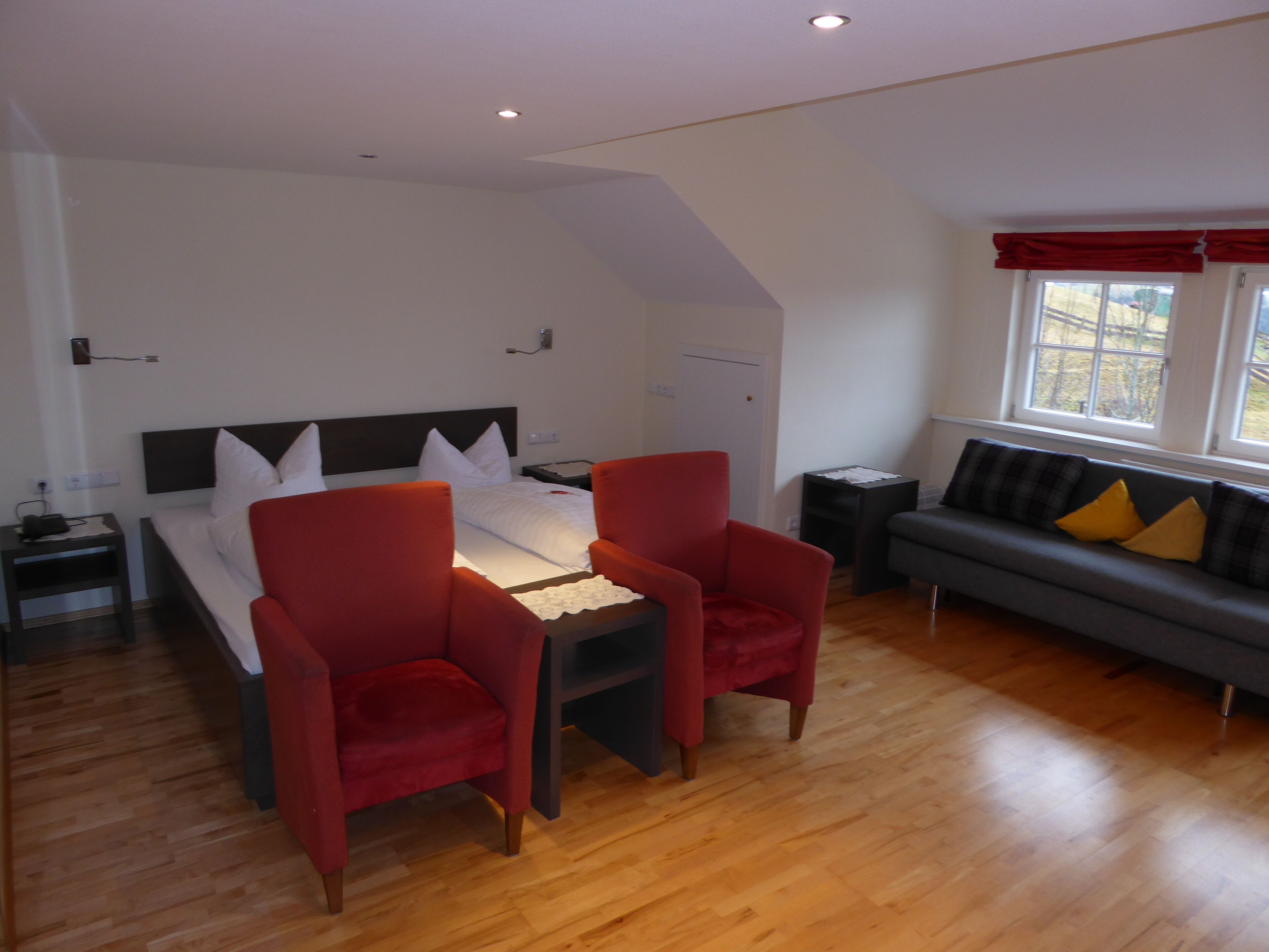 Une chambre d’hôtel moderne avec un grand lit, deux fauteuils rouges, un canapé gris avec des coussins jaunes, un plancher en bois et une fenêtre avec des rideaux rouges.