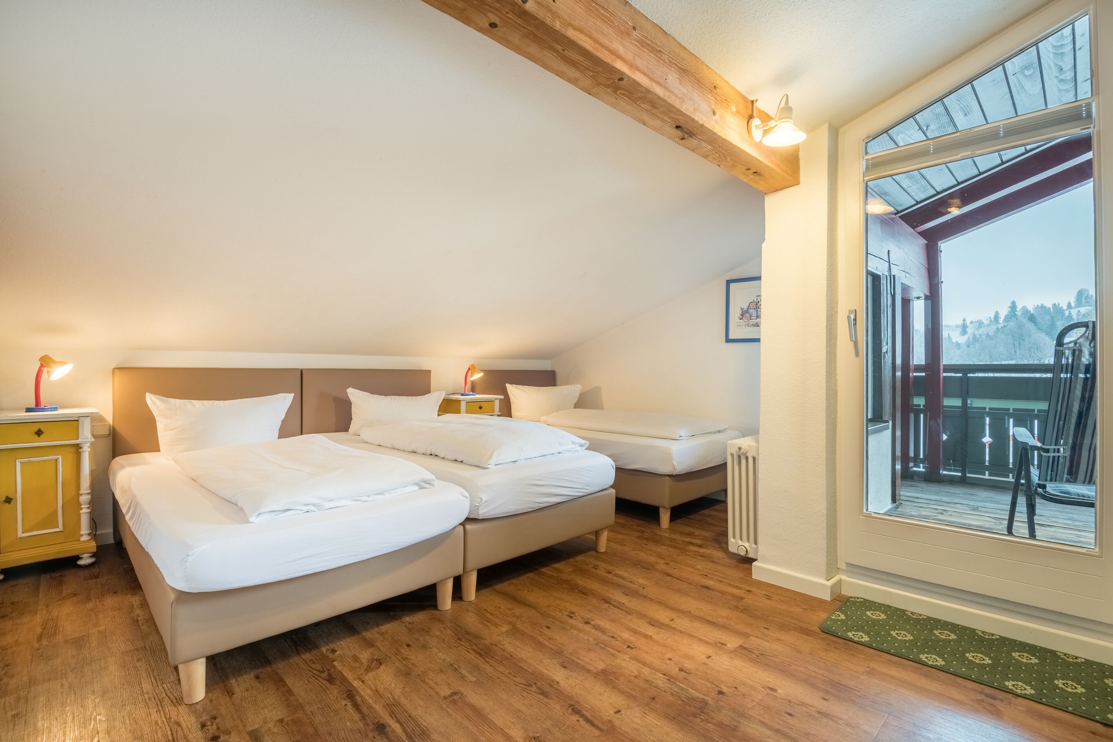 A bedroom with two beds, wooden flooring, a large window with a view, and a wooden ceiling beam.