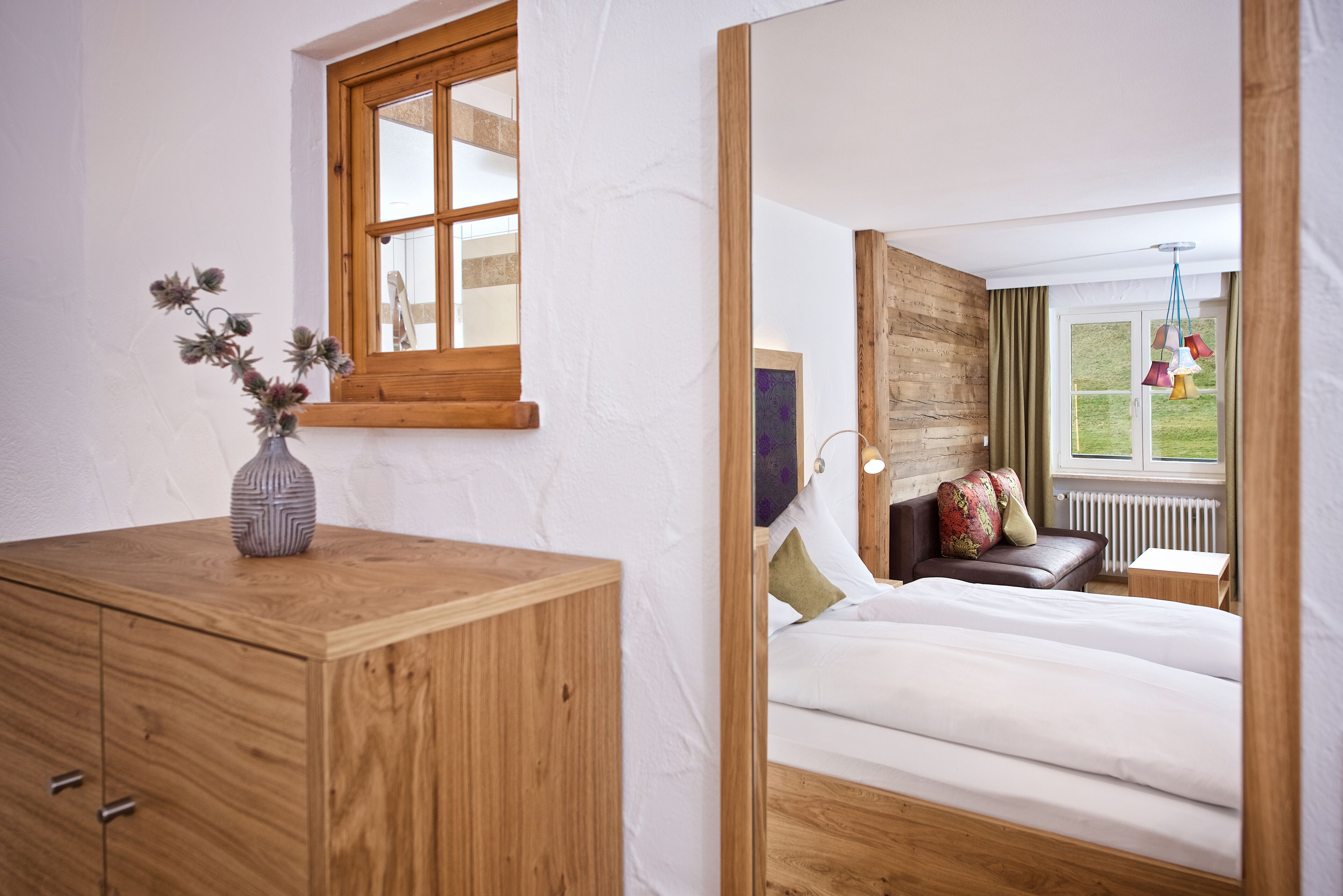 A bedroom with a wooden dresser, a window with a view, a bed with pillows, and a hanging light fixture.