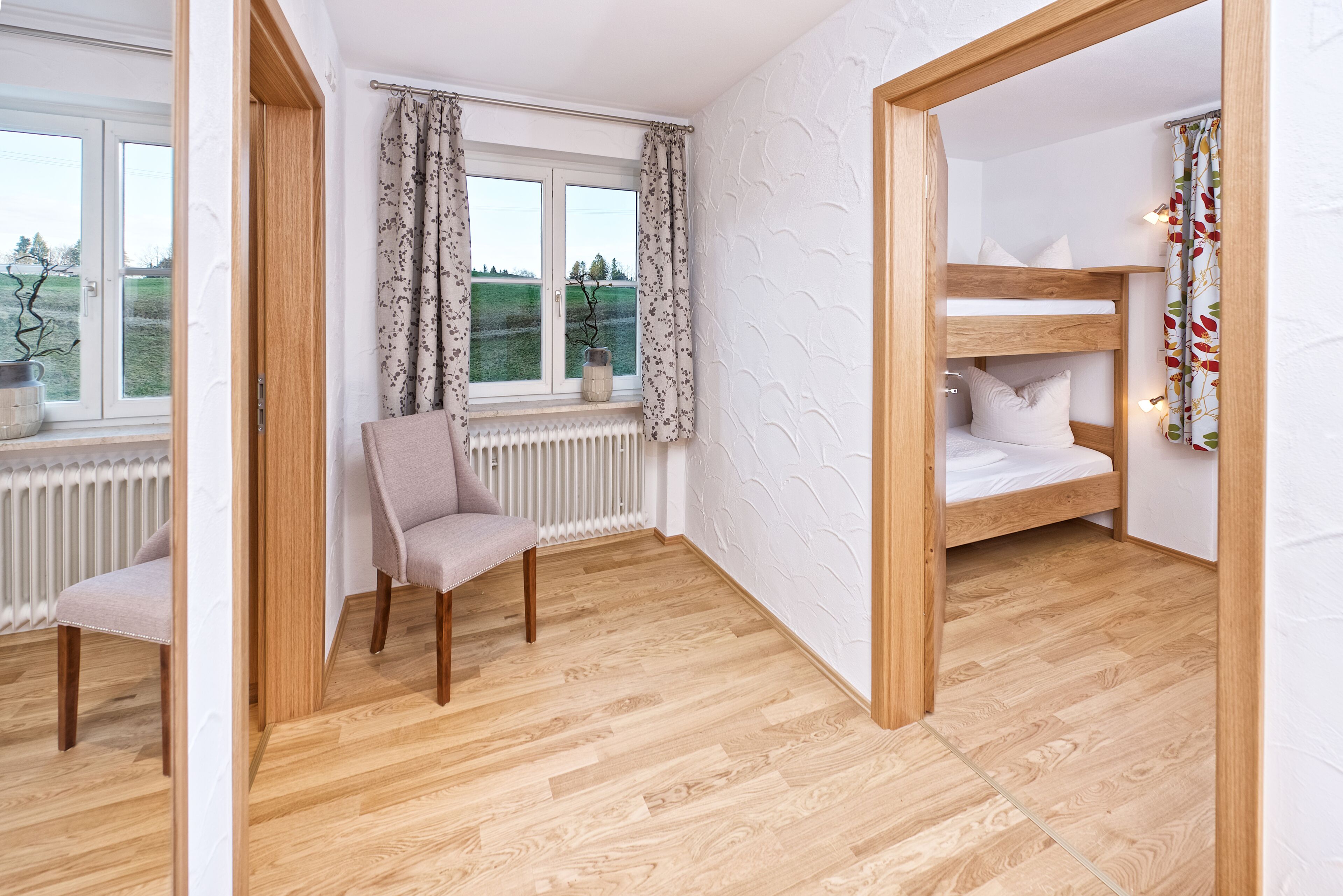 A room with a wooden floor, a chair, a window with curtains, and a bunk bed with pillows.