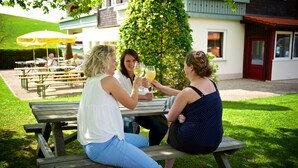 Tres personas sentadas en una mesa de picnic de madera al aire libre, con un edificio y un árbol de fondo.