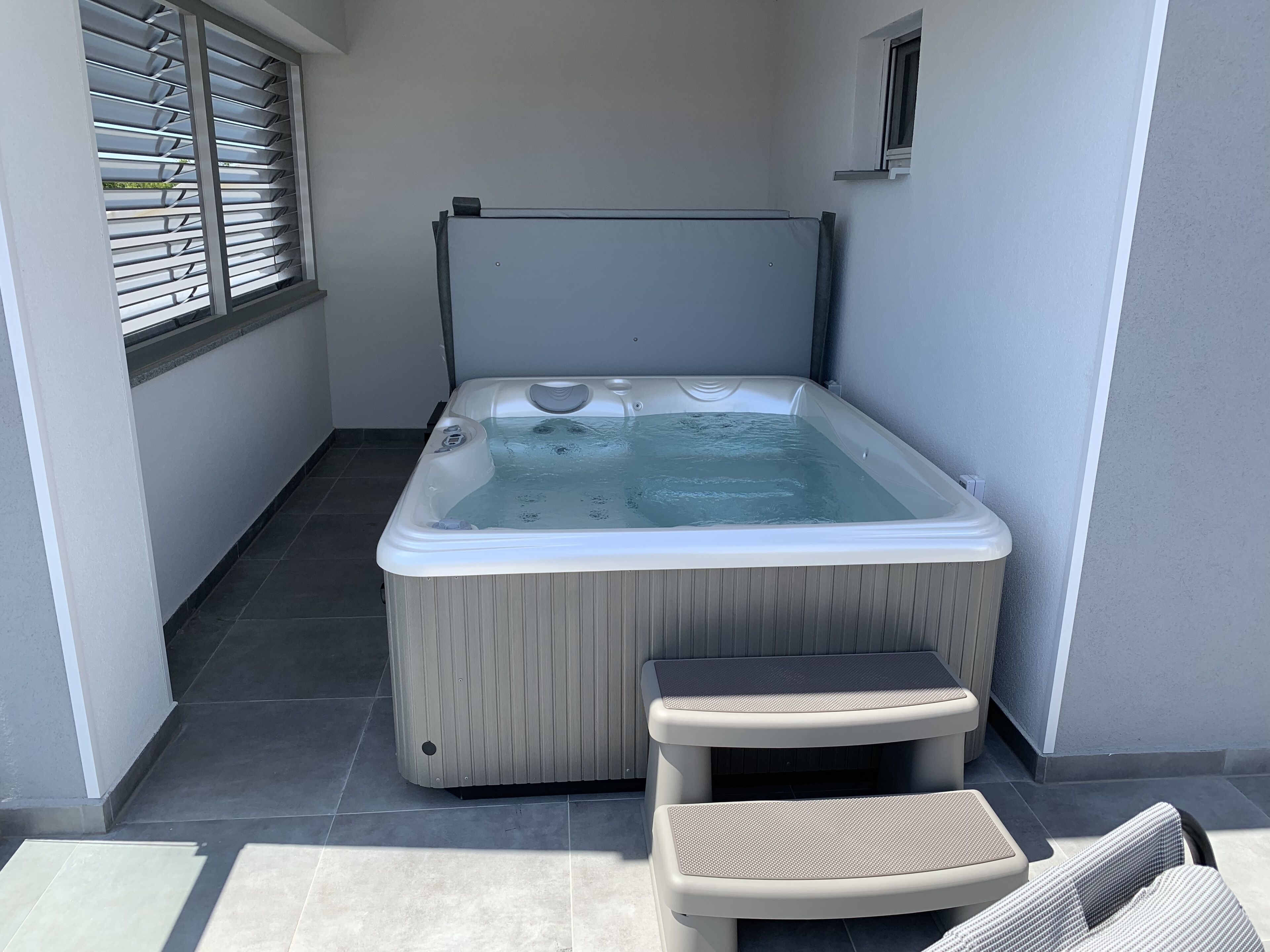A hot tub with a control panel, a small table, and a chair in a tiled outdoor area.