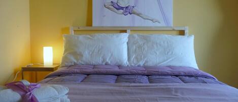 A neatly made bed with a purple blanket and pillows, a white headboard, and a painting of a dancer on the wall.
