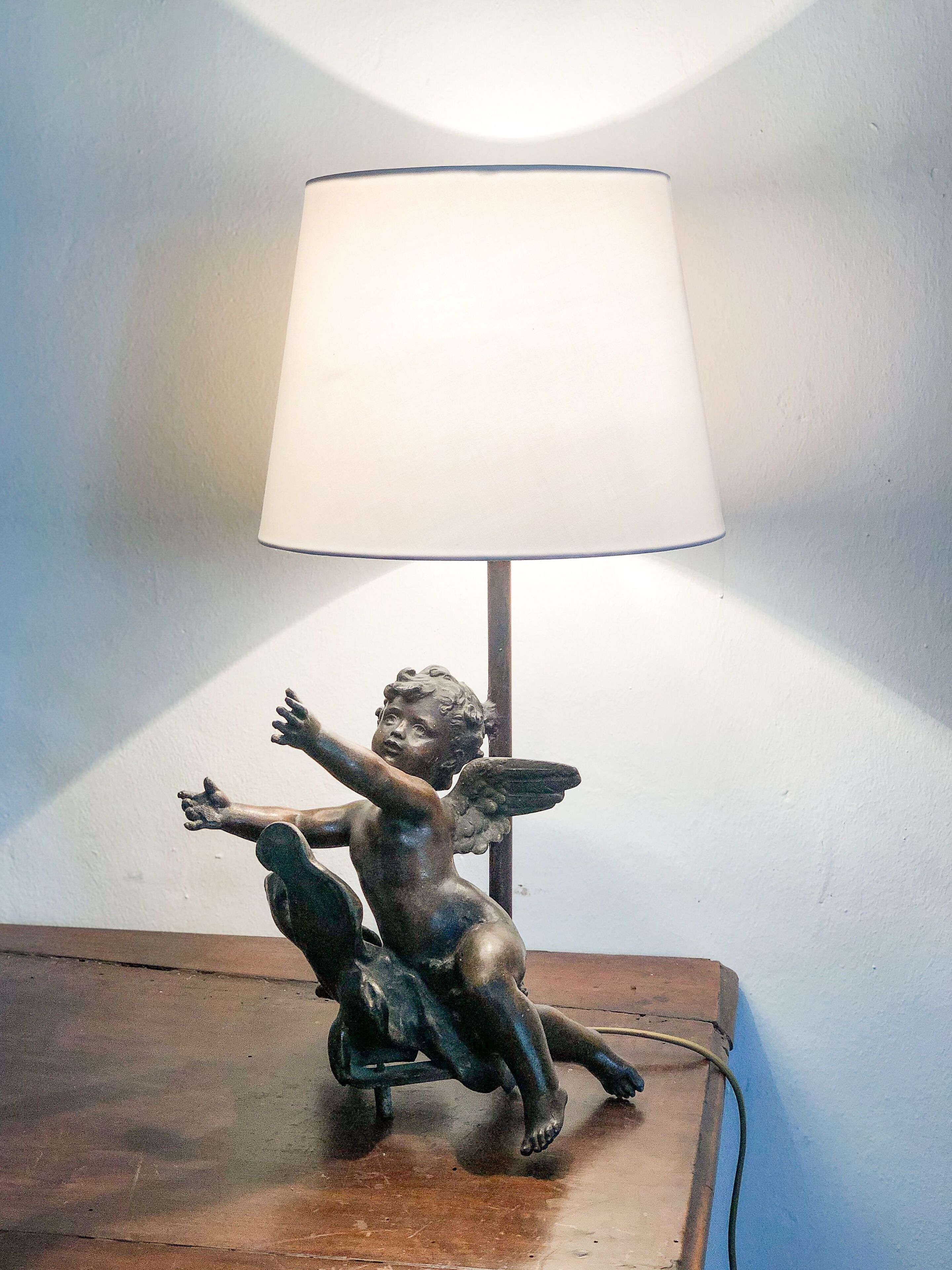 A statue of a cherub with outstretched arms on a wooden table, under a lamp with a white shade.