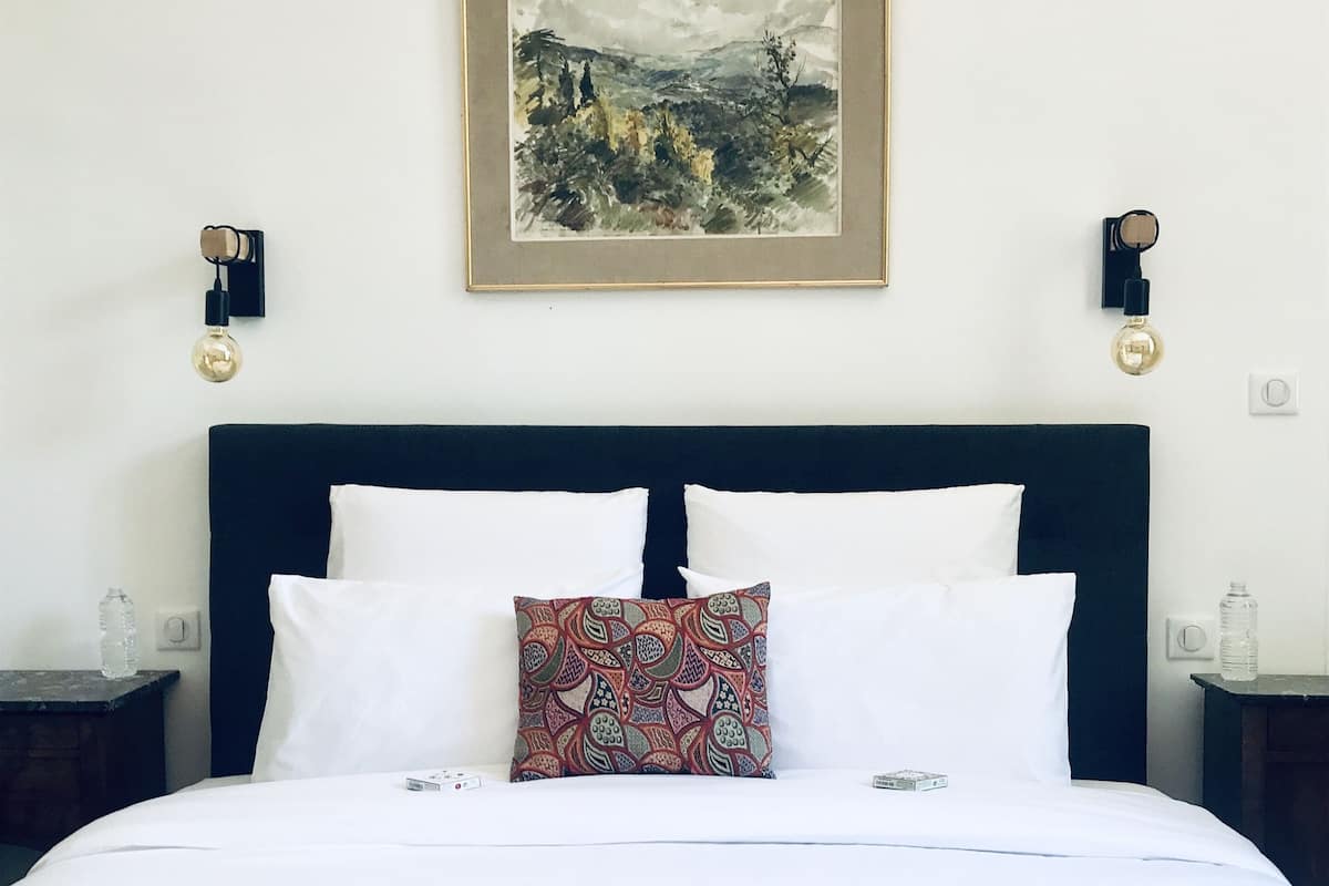 Un lit bien fait, agrémenté d’un coussin à motifs, avec un tableau représentant un paysage encadré au-dessus, et deux appliques murales.