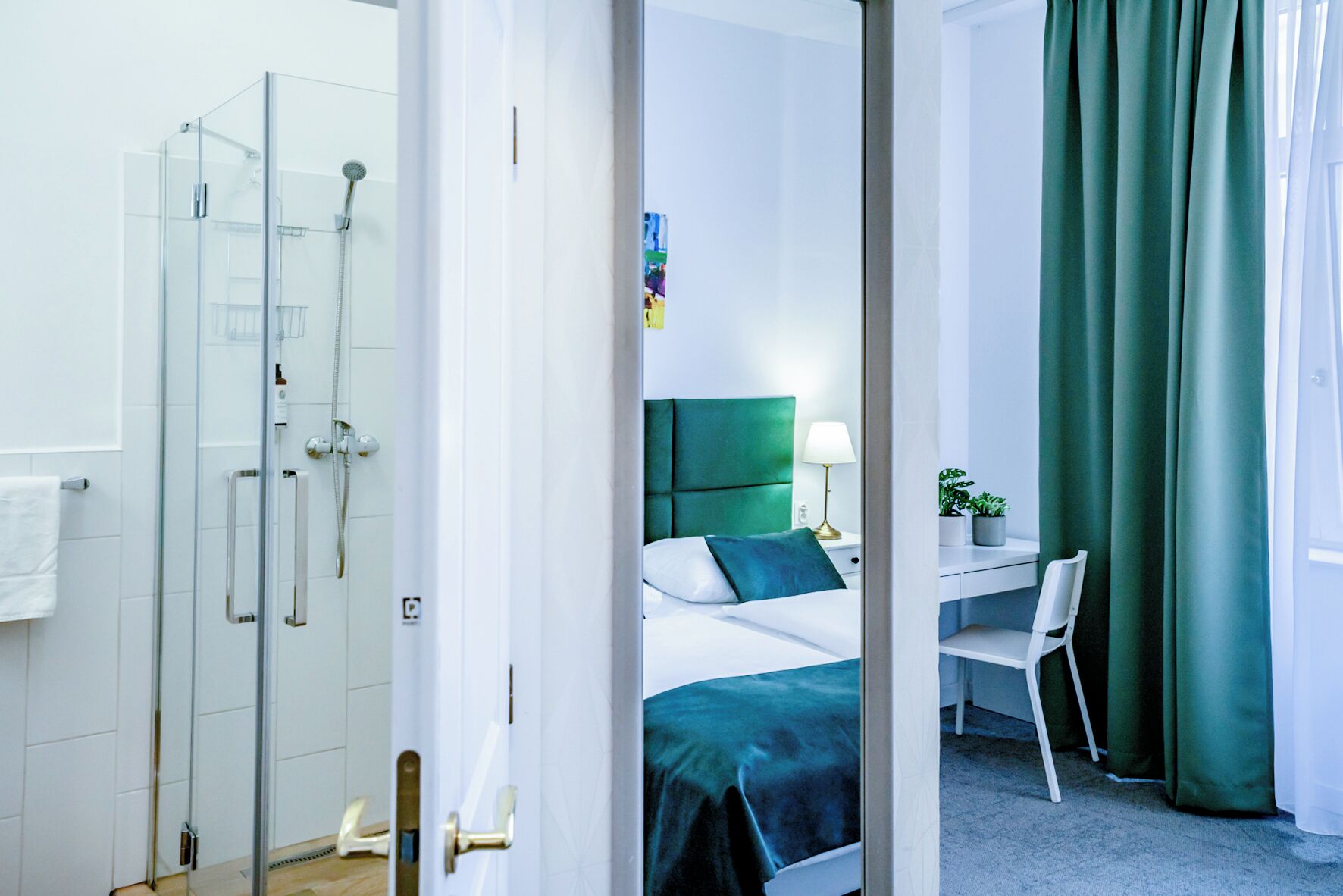 Une chambre d’hôtel avec une douche en verre, un lit avec une tête de lit verte, un bureau avec une chaise et un rideau vert.