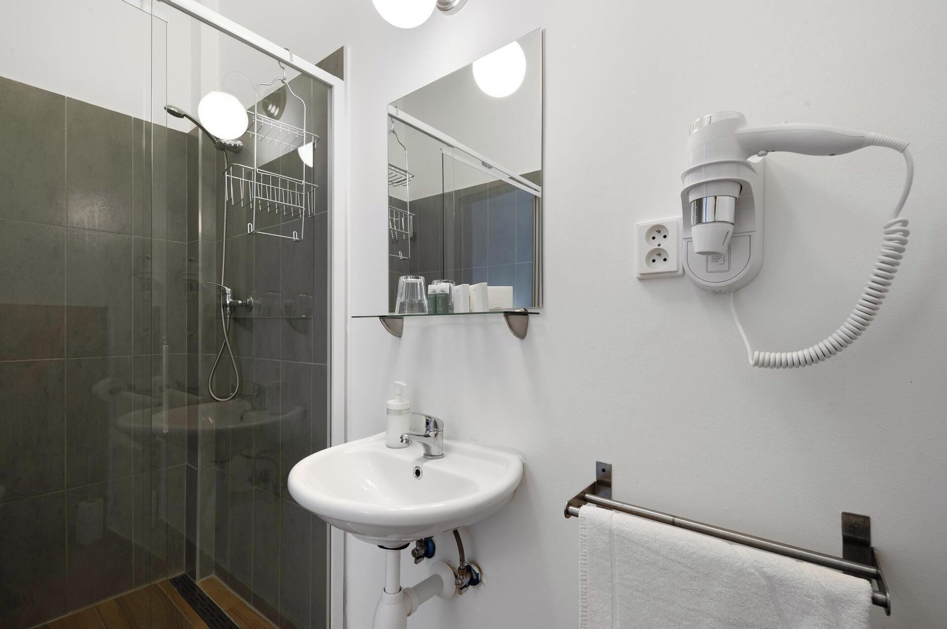 Un baño moderno con cabina de ducha de vidrio, un lavamanos blanco, un espejo y un secador de pelo montado en la pared.
