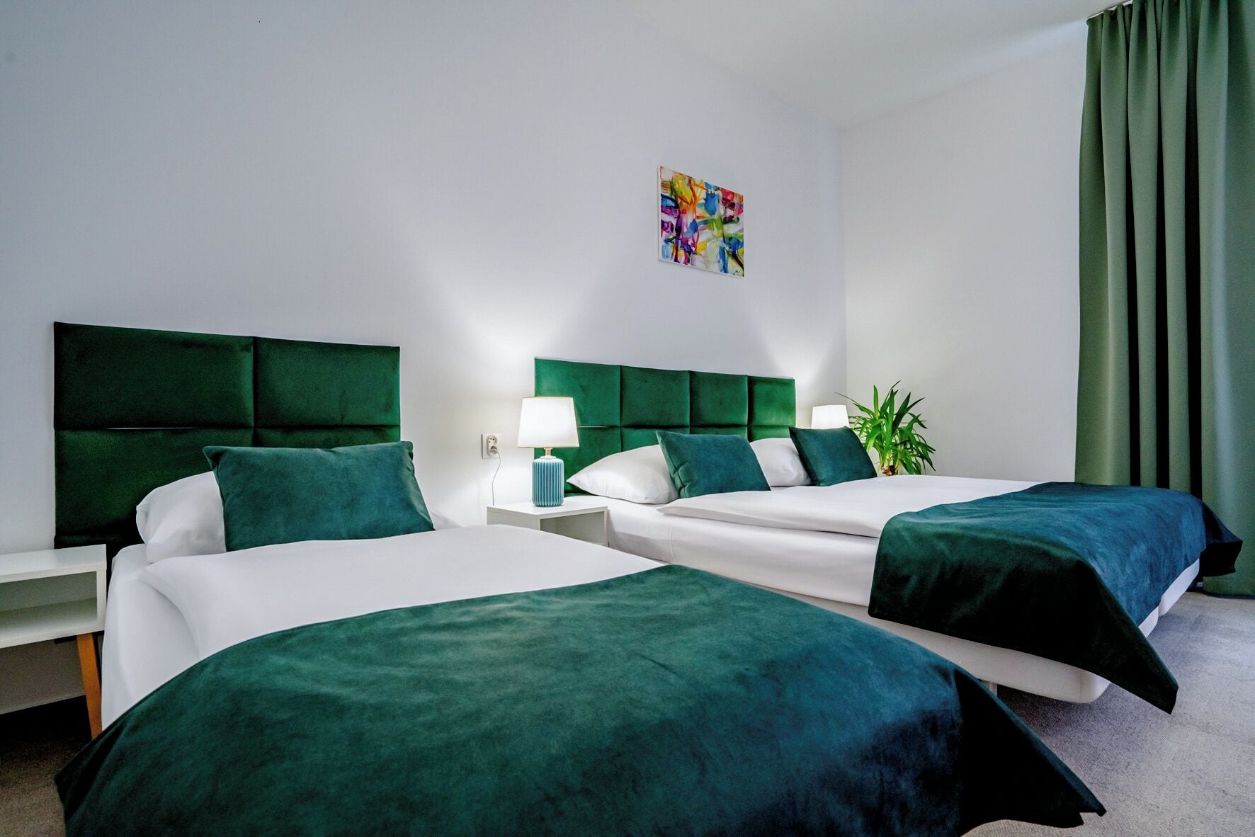 A hotel room with two beds, each with green headboards and white bedding. There is a small bedside table with a lamp and a plant on the right side.