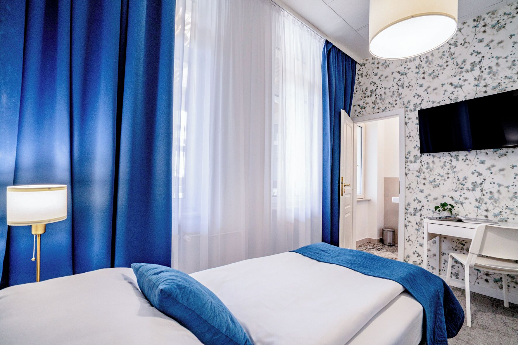 Une chambre d’hôtel avec un lit, un bureau, une chaise, une télévision et une fenêtre avec des rideaux.
