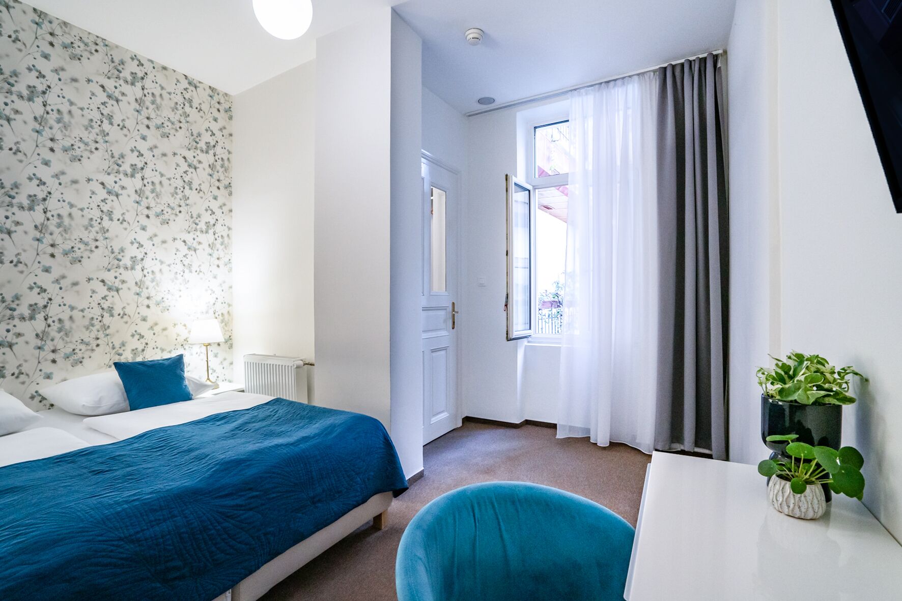 A hotel room with a bed, a desk, a chair, a radiator, a window with curtains, and a potted plant.