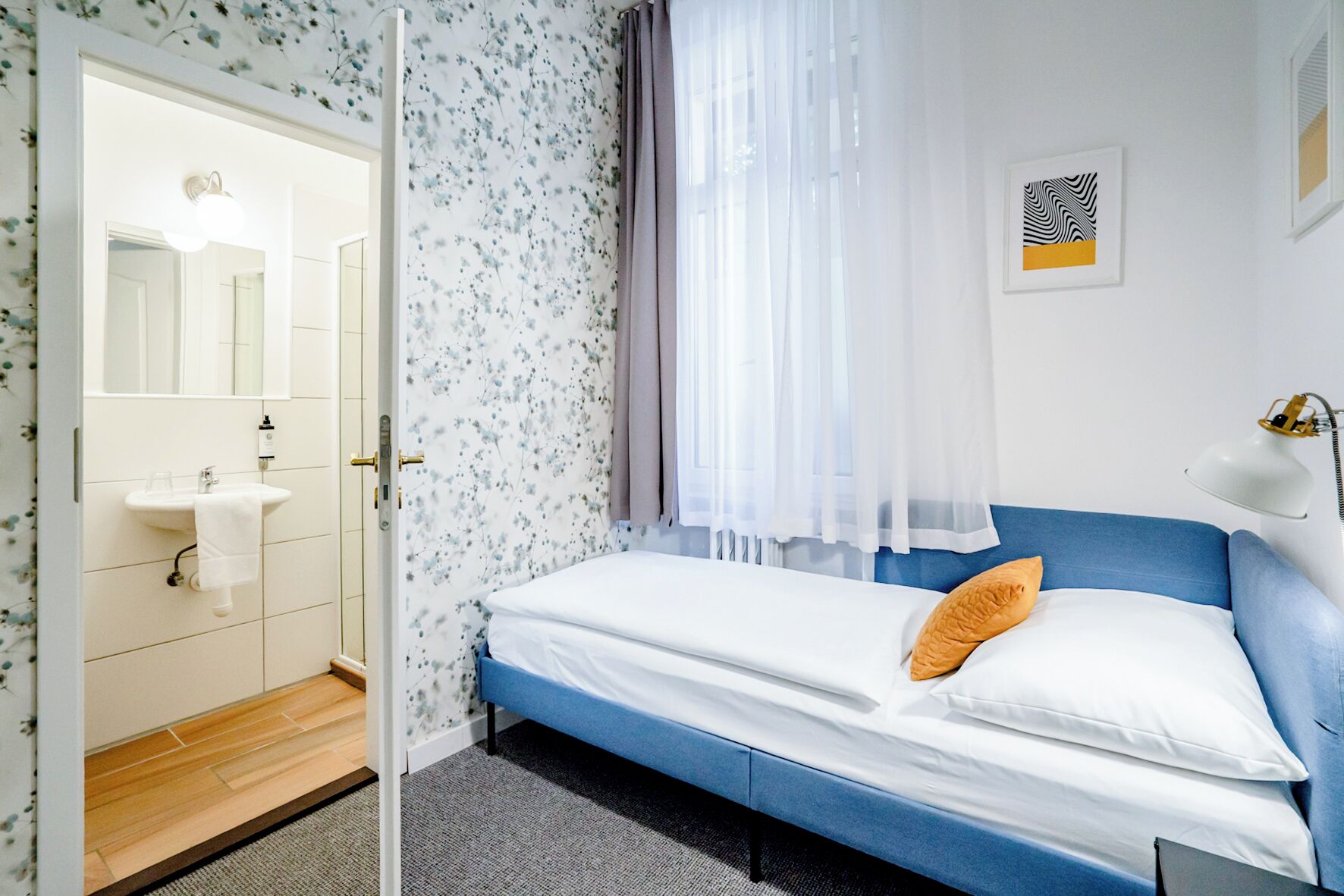 A bedroom with a bed, a bedside lamp, a window with curtains, a sink with a towel, and a mirror.