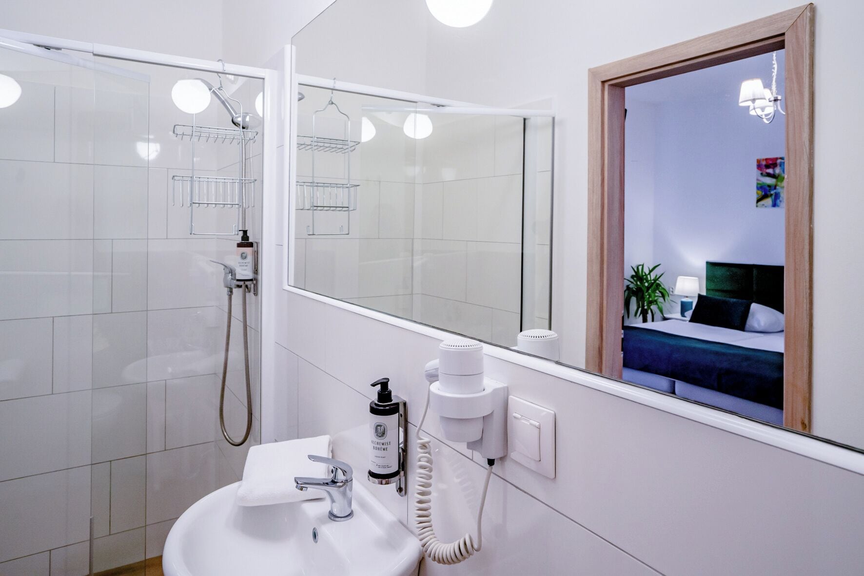 A modern bathroom with a glass shower enclosure, a white sink, and a mirror reflecting a bedroom with a bed and a nightstand.