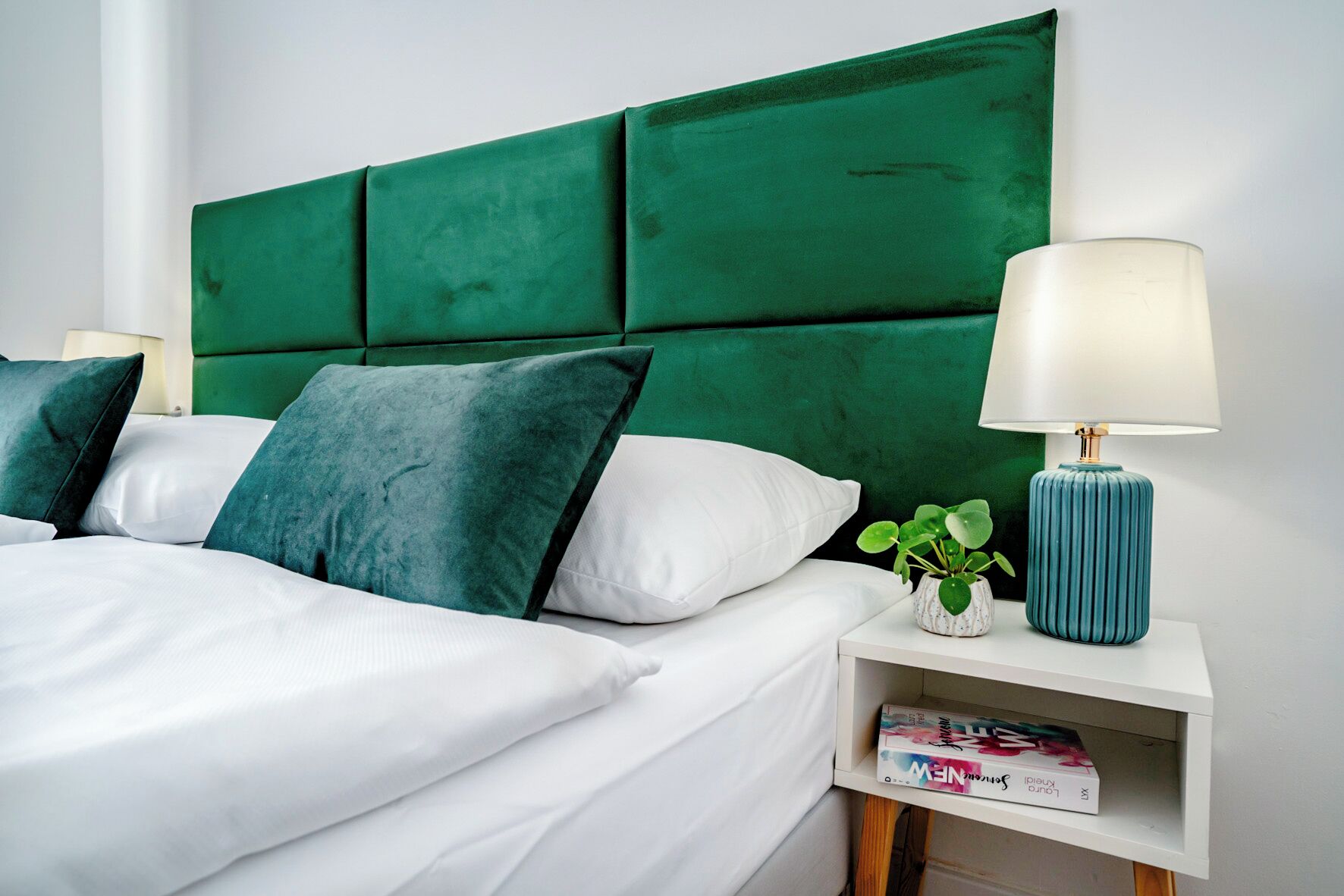 Una cama bien hecha con ropa de cama blanca y verde, un cabecero tapizado en verde y una mesita de noche con una lámpara y una planta pequeña.