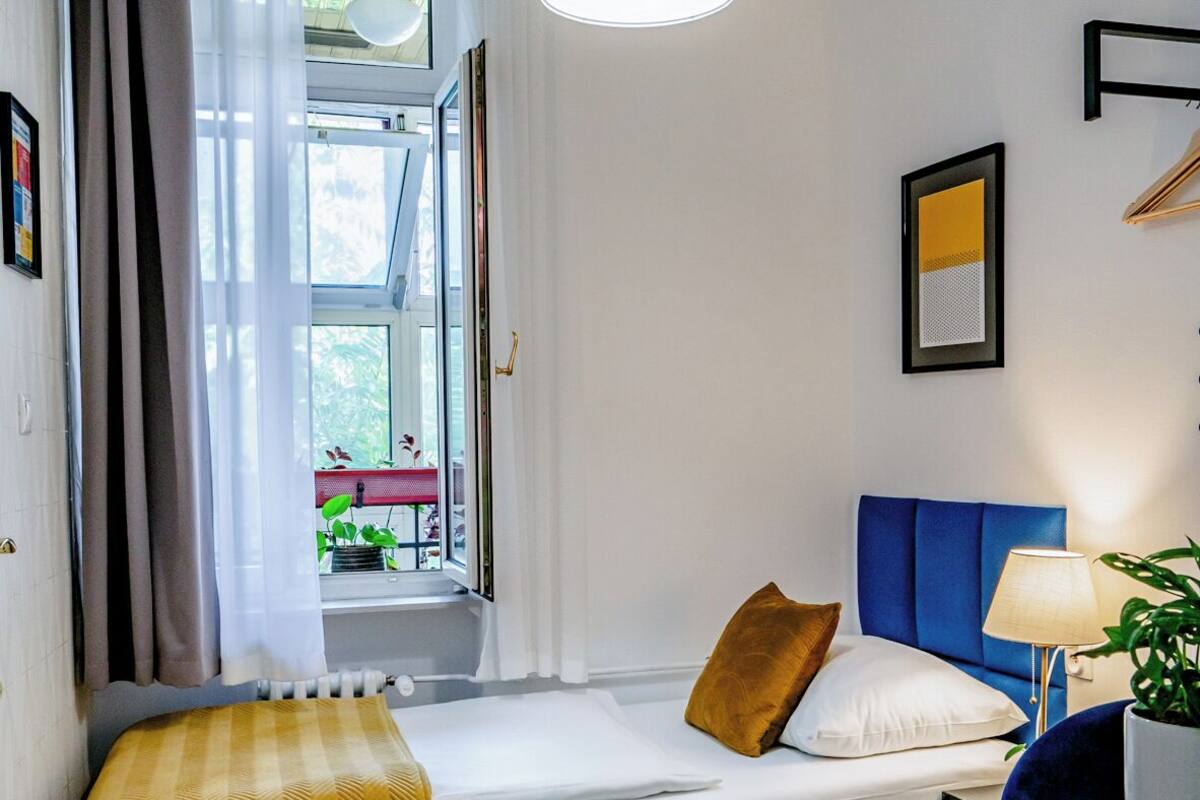 A compact room with a bed, a window with curtains, a hanging light fixture, a chair, and a table with a plant.