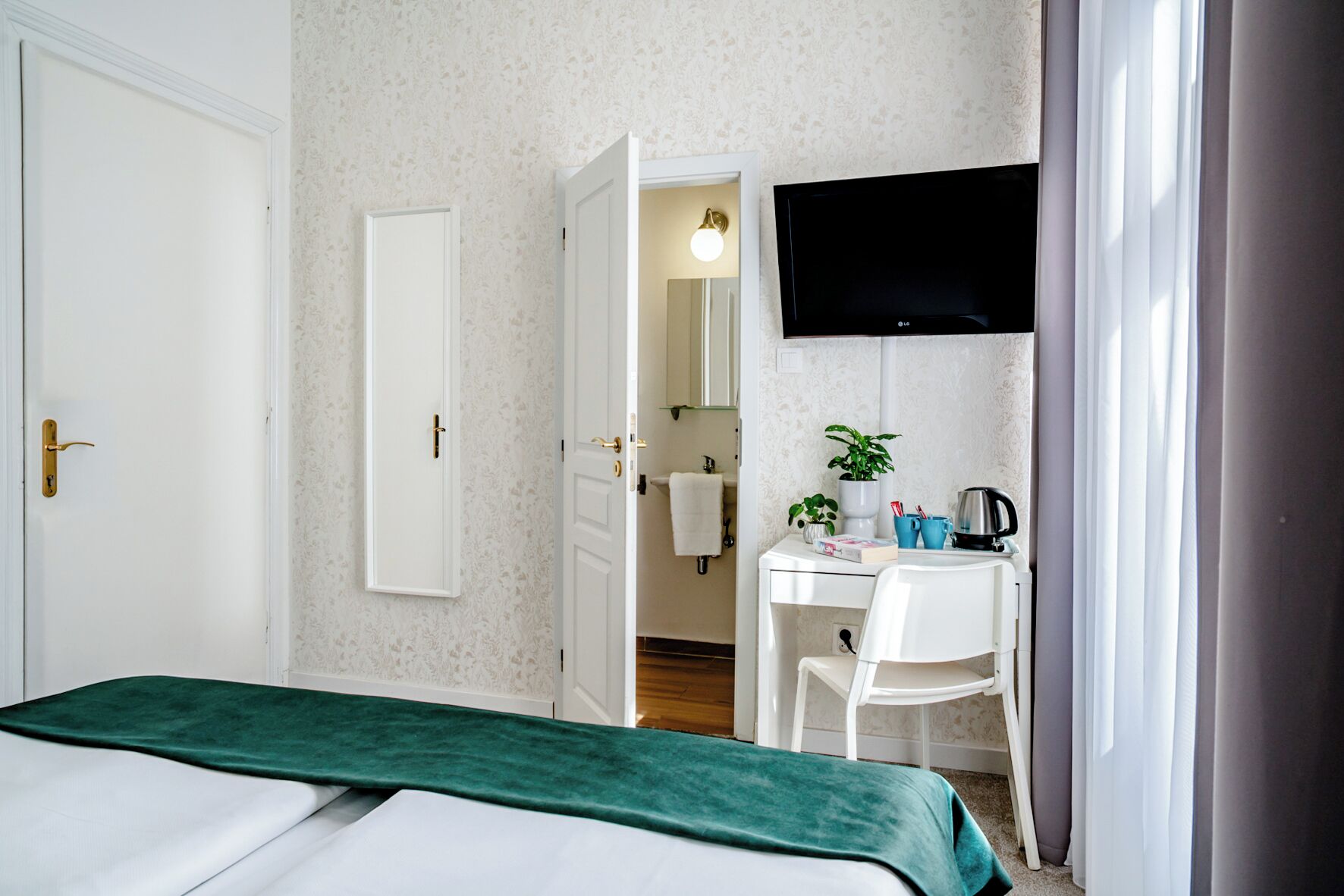 A hotel room with a bed, a desk, a chair, a TV, and a bathroom visible through an open door.