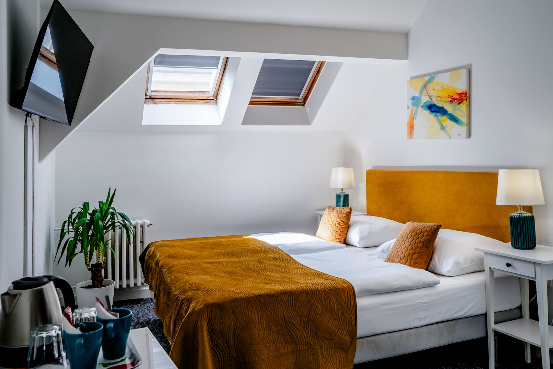 A hotel room with a bed, two bedside tables, a TV, a plant, and a painting on the wall.