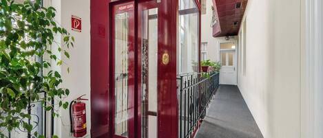 Une porte rouge avec des panneaux de verre, un extincteur et une rambarde métallique le long d’un couloir.