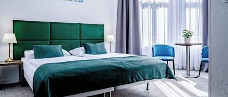 A hotel room with a green upholstered bed, two bedside tables with lamps, a small round table with a potted plant, and a blue chair.