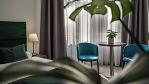 A hotel room with a bed, a nightstand, a lamp, two chairs, a small table with a plant, and a window with curtains. - Elisabeth Old Town (Bratislava)
