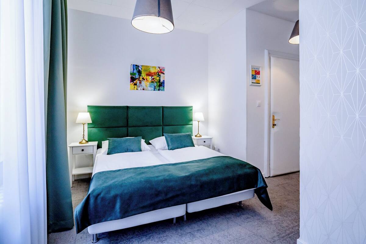 A modern hotel room with a large bed, green headboard, two bedside tables with lamps, and a colorful abstract painting on the wall.