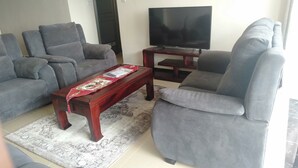 A living room with grey sofas, a wooden coffee table with a red table runner, a flat-screen TV on a wooden stand, and a patterned rug on the floor. - S. Lucie Mango Apartment (Nairobi)