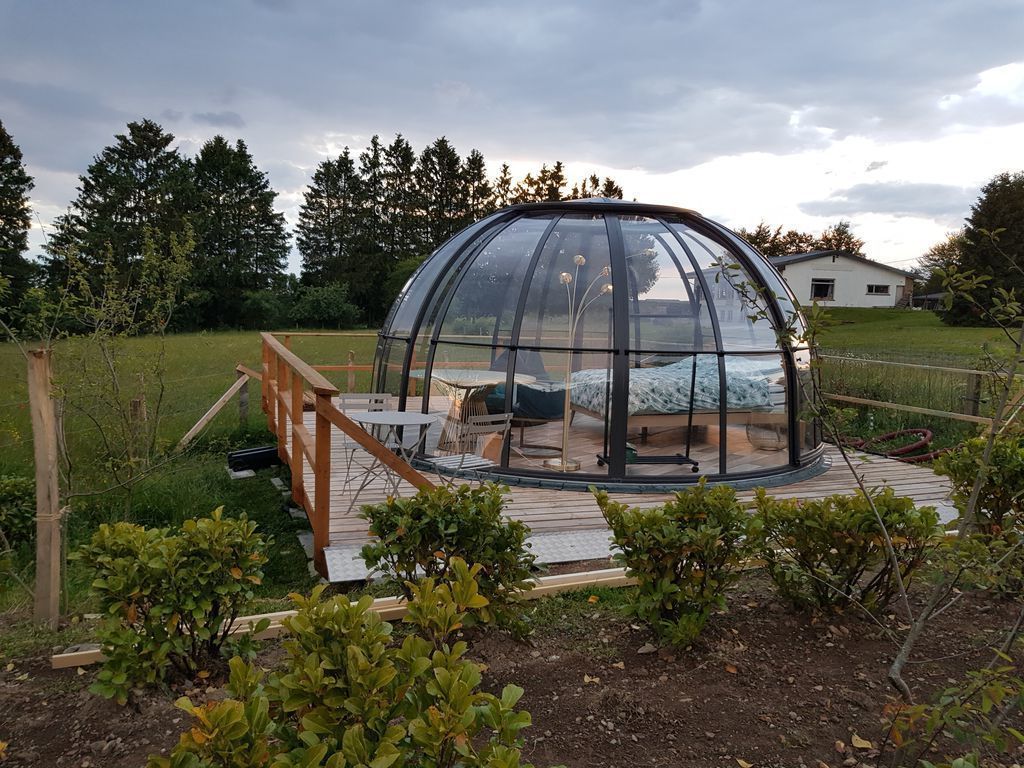 Een koepelvormige glazen constructie met een houten terras en zitgedeelte, omgeven door groen en bomen.