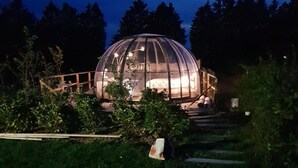 A dome-shaped glass structure with a bed inside, surrounded by a wooden deck and steps leading up to it. - La bulle au bois dormant d'ovifat (Waimes)