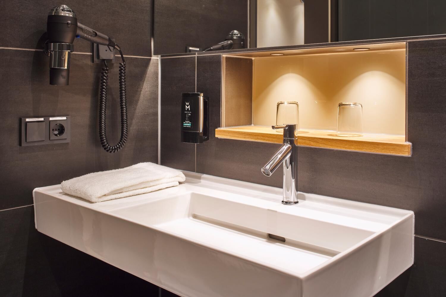 A modern bathroom with a white sink, a handheld hair dryer, and a mirror with a wooden shelf.