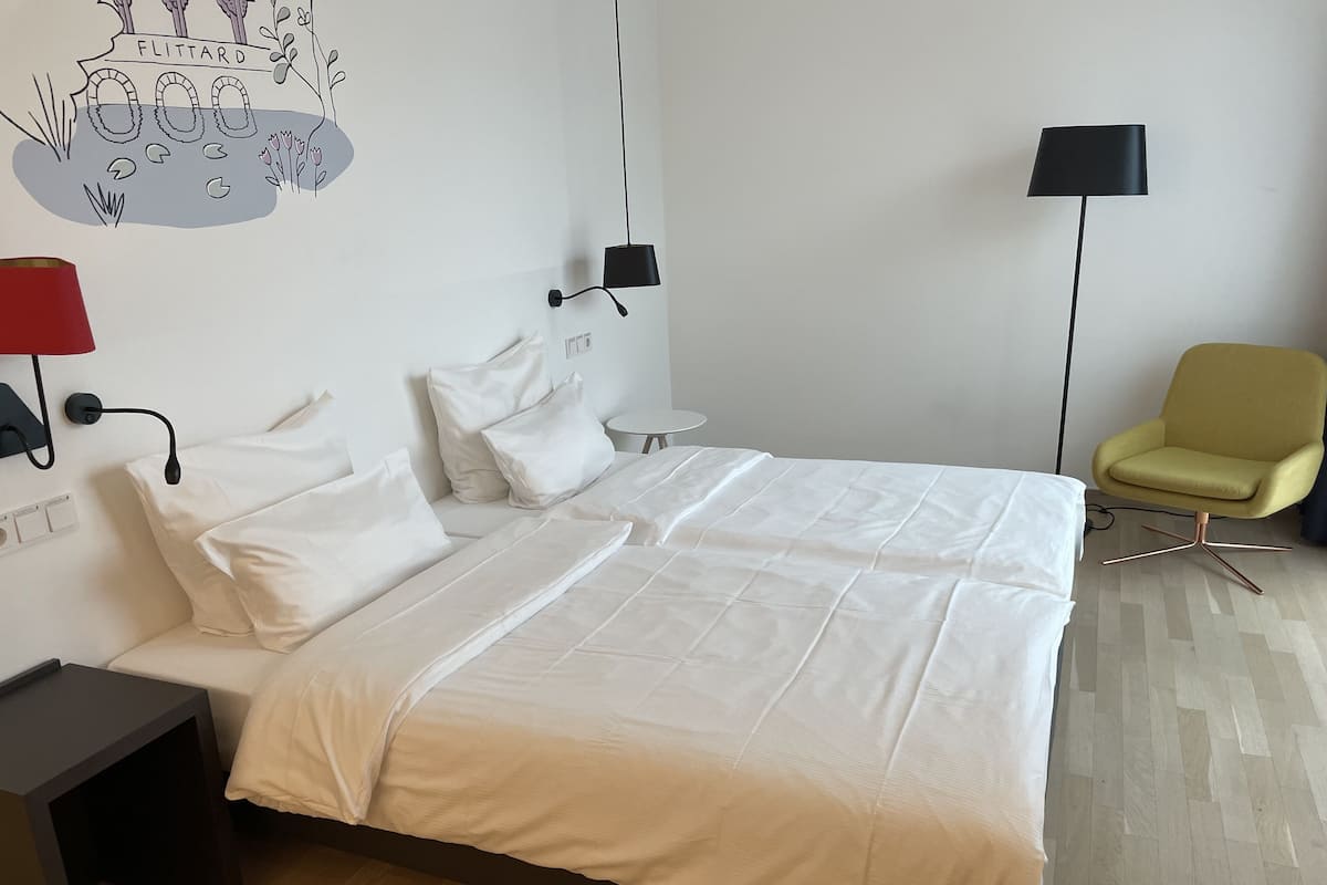 Een hotelkamer met een tweepersoonsbed, een klein nachtkastje, een stoel, een lamp en een decoratie aan de muur.
