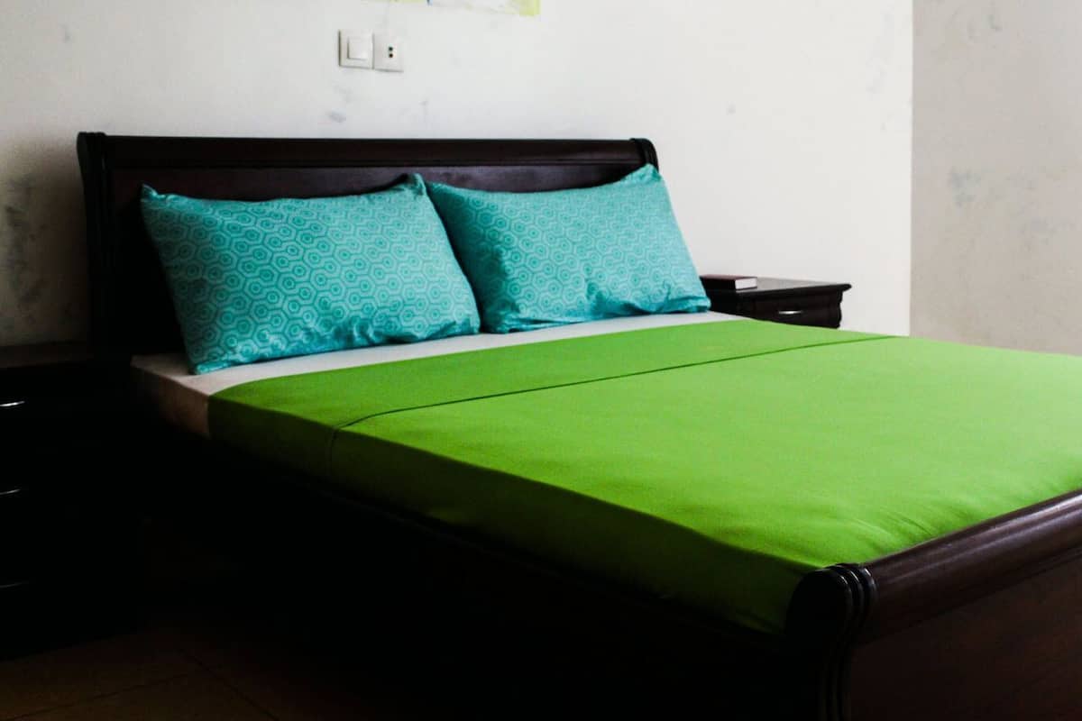 A bed with a green bedspread and two teal pillows, a wooden headboard, and a nightstand with a lamp.