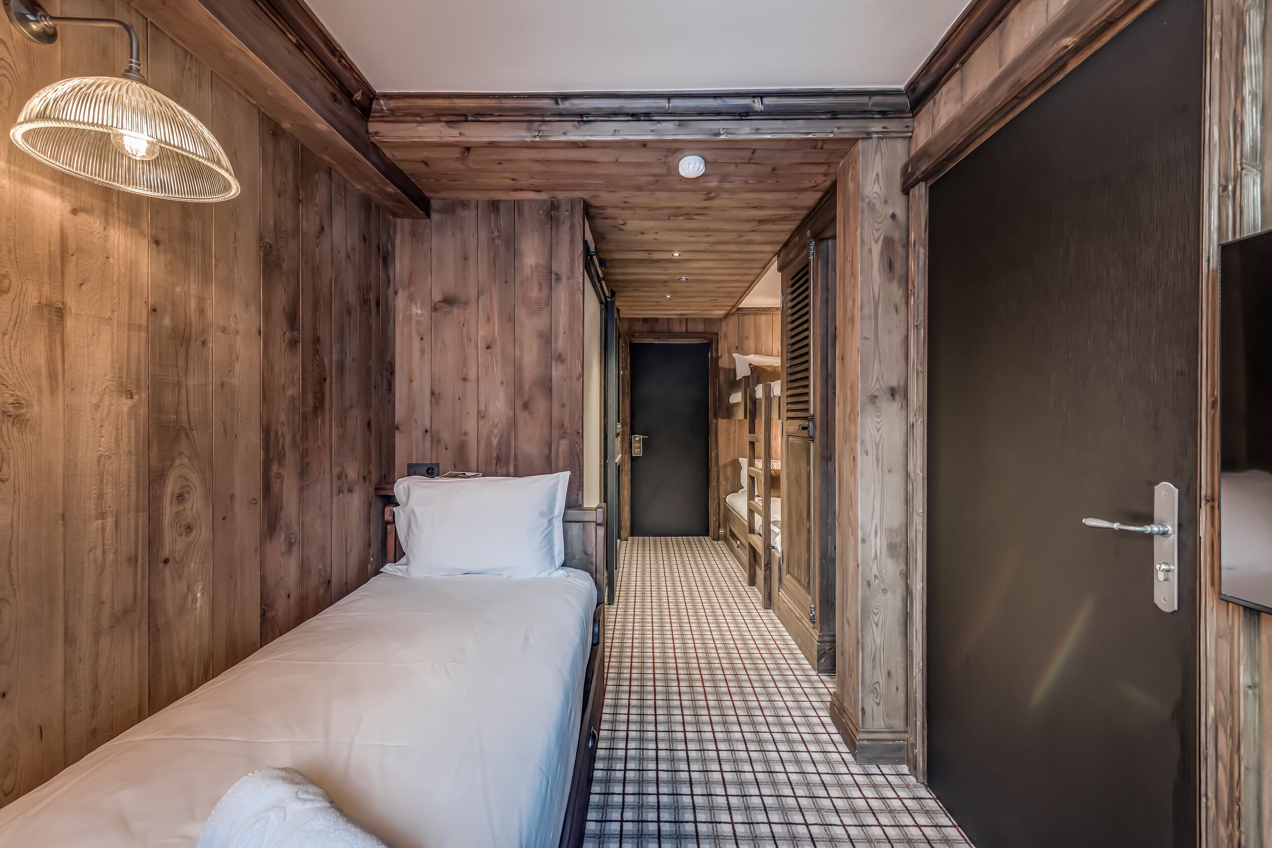 Un couloir étroit aux murs en bois, un lit recouvert de draps blancs et un sol carrelé.