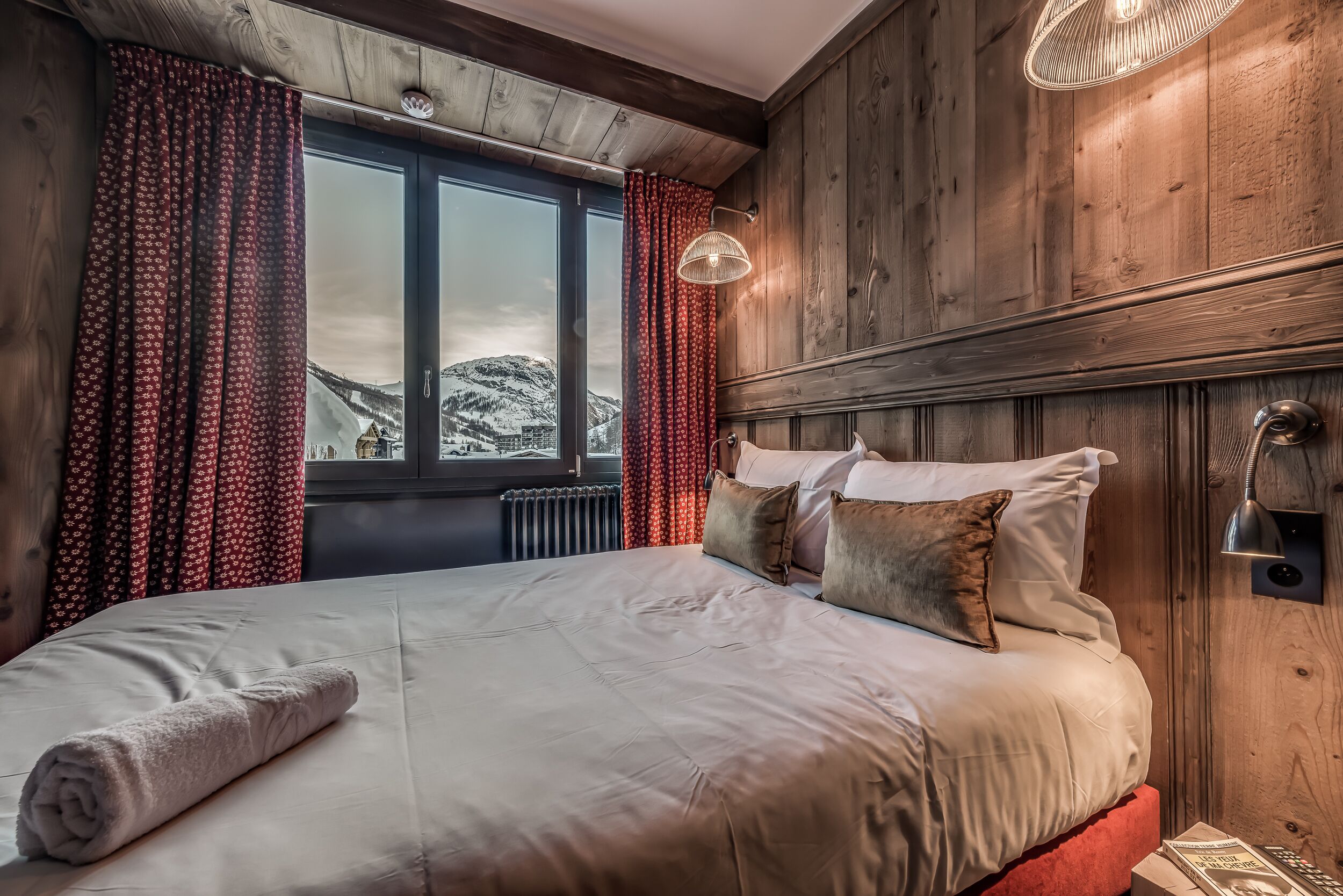 A cozy bedroom with a large bed, wooden paneling, a view of snow-capped mountains, and a window with red curtains.