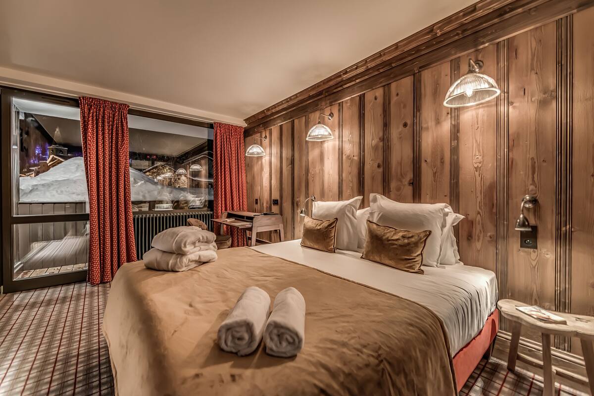 A hotel room with a large bed, wooden panel walls, and a view of a snowy landscape through a glass door.