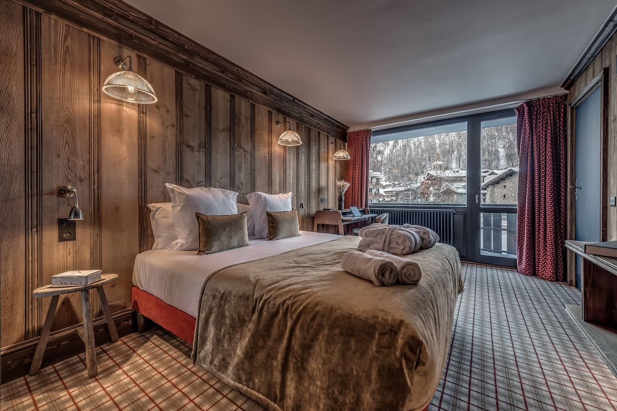 A cozy hotel room with a large bed, wooden paneling, a view of a snowy landscape, and a small table with a chair.