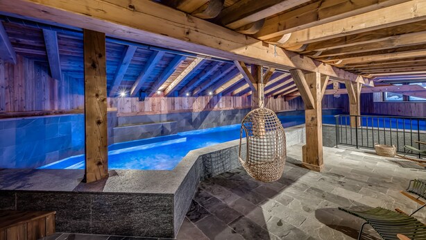 Una piscina interna con travi in legno, pavimento in pietra e una sedia in vimini sospesa.