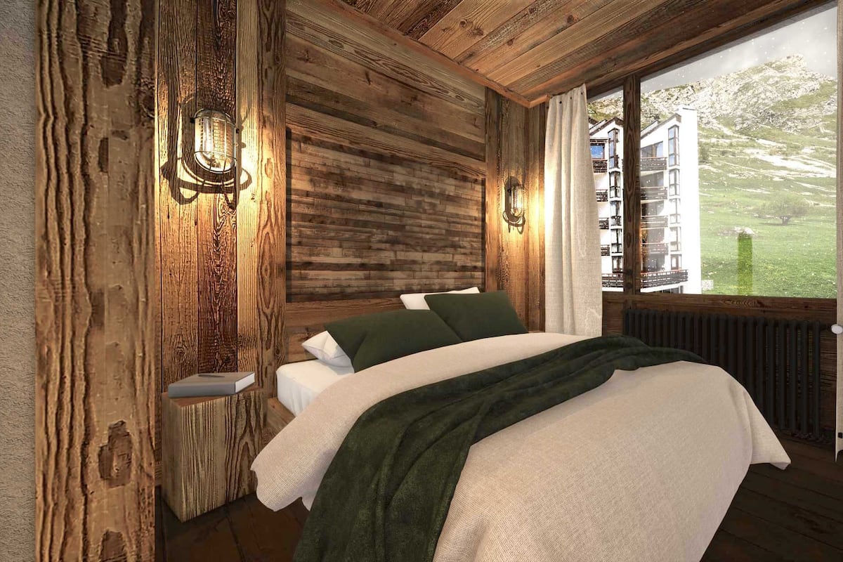 A bedroom with a wooden ceiling, a bed with white and green bedding, a nightstand, a wall-mounted light, and a window with curtains.