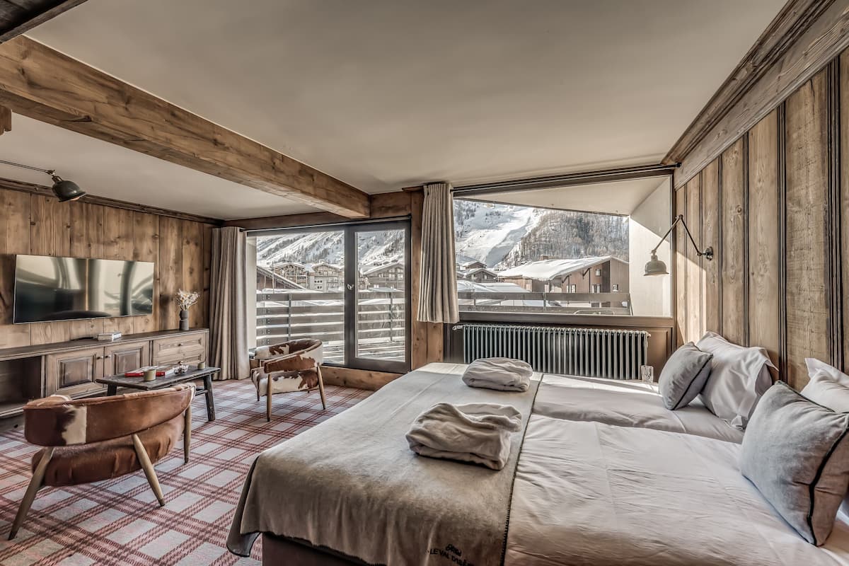 A modern hotel room with a large bed, a TV, a wooden desk, and a view of a snowy mountain.