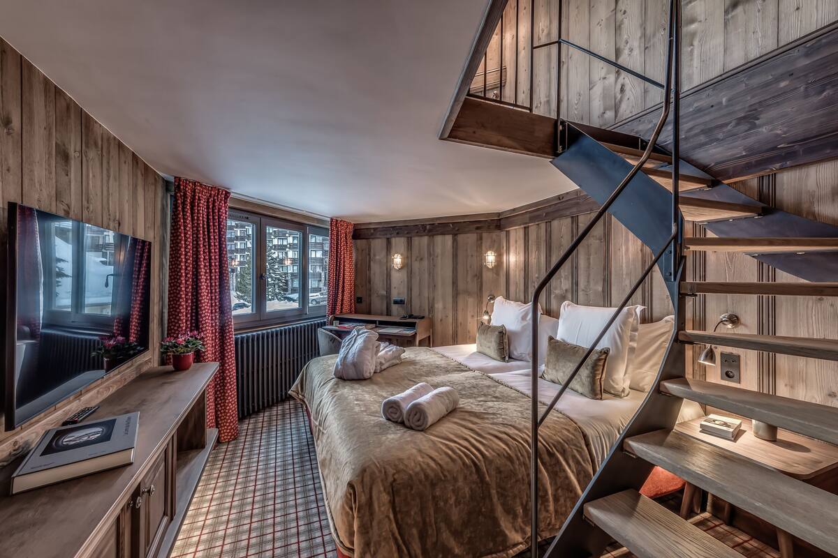 A modern hotel room with a large bed, a TV, a desk, and a wooden staircase.