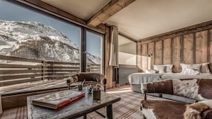 Una stanza accogliente con un'ampia finestra che si affaccia su una montagna innevata, un tavolo di legno con un libro, un divano con una coperta di pelliccia e un letto con biancheria bianca.
