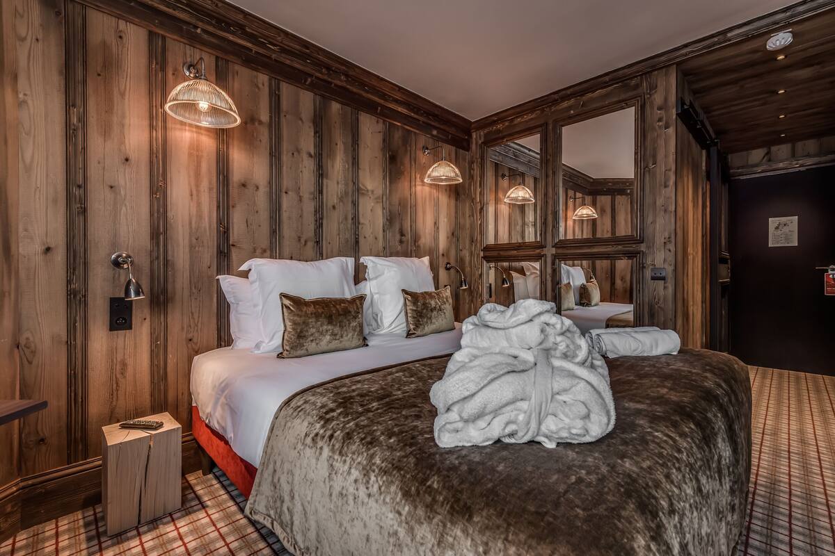 A wooden-paneled bedroom with a large bed, two bedside lamps, a mirror, and a small bedside table with a remote control.