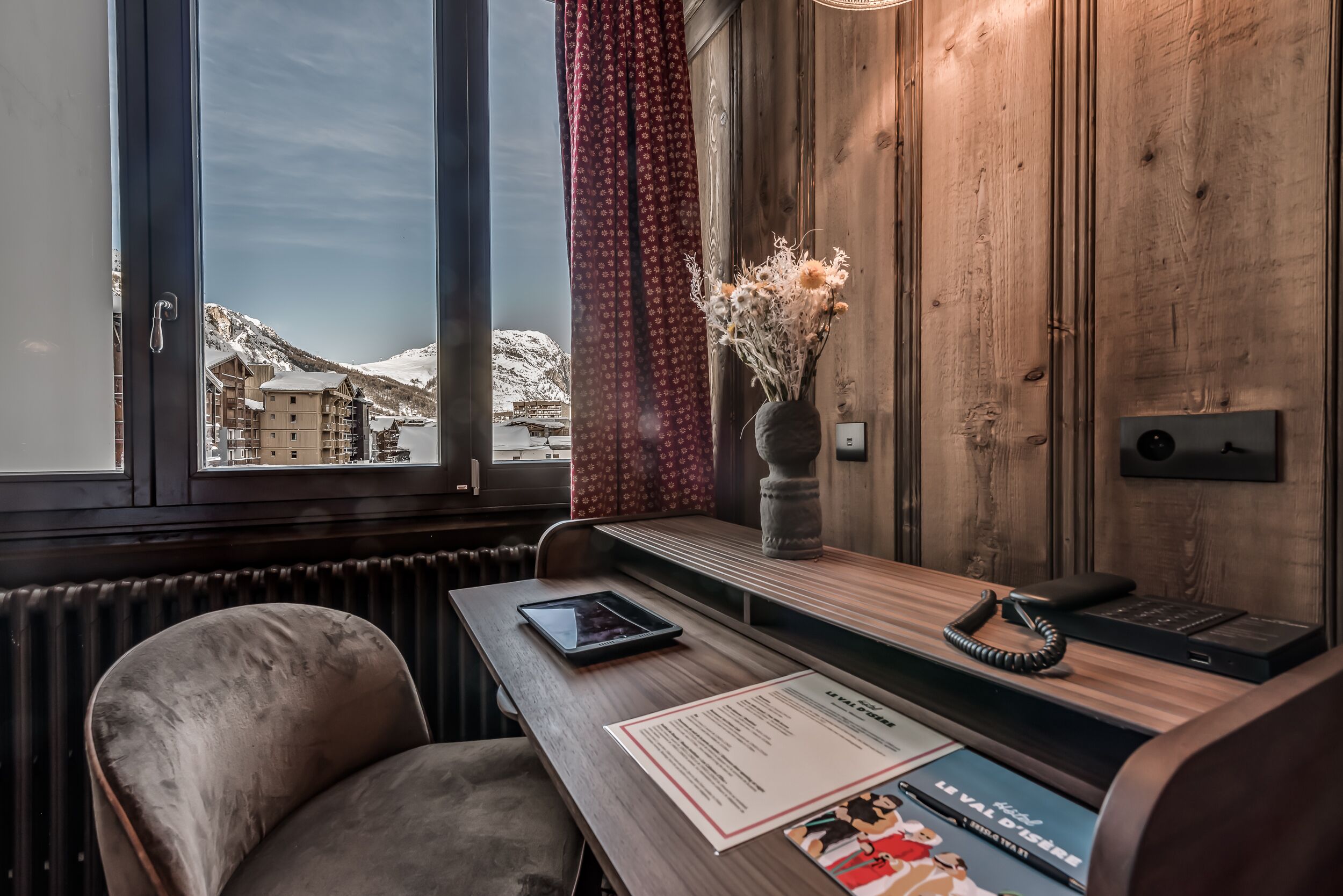 Een hotelkamer met een houten bureau, een tablet, een telefoon en een vaas met bloemen. Een raam biedt uitzicht op besneeuwde bergen.
