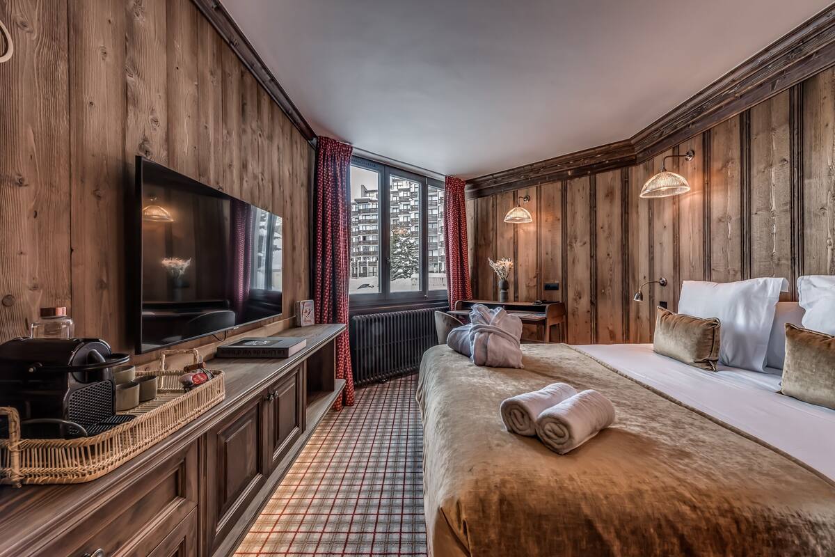 A cozy room with wooden walls, a large bed with a fur throw, a flat-screen TV, and a window with red curtains.