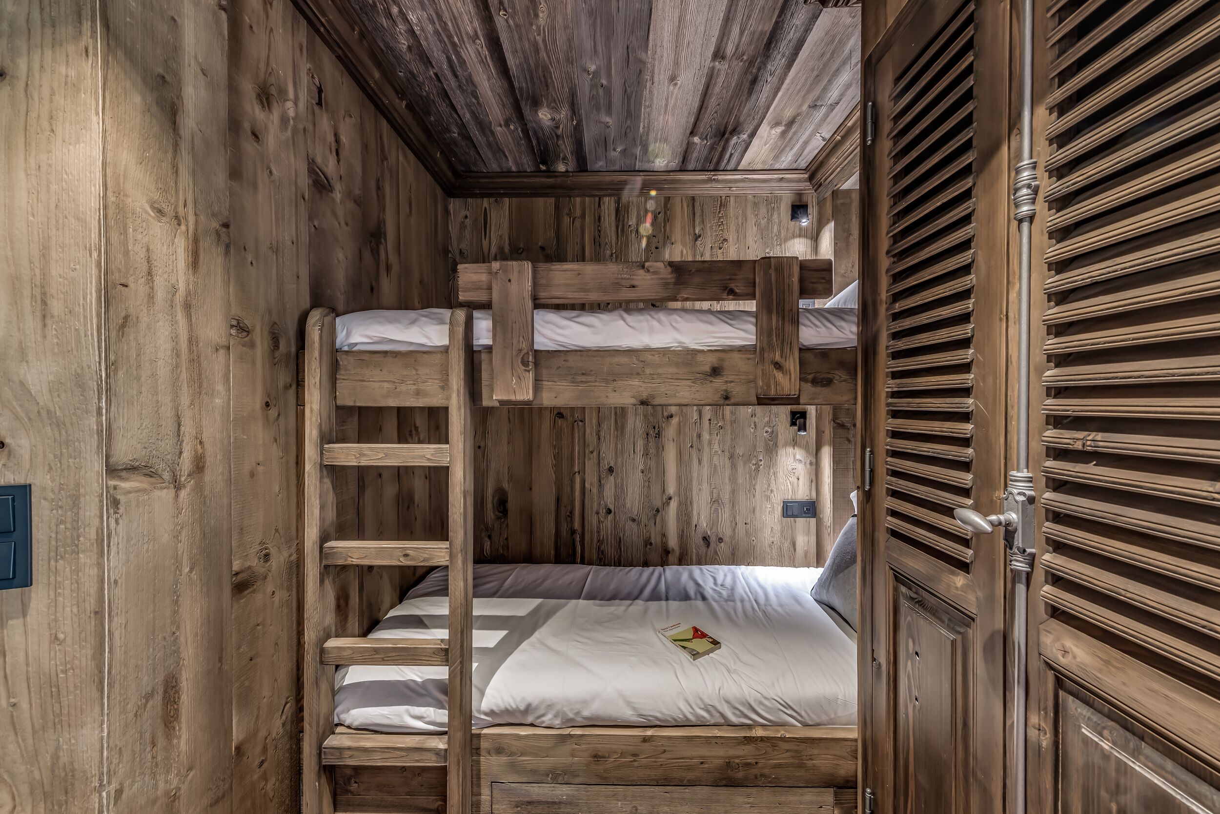Ein Zimmer mit einem hölzernen Etagenbett, einer Leiter, einer Tür mit Griff und einer kleinen Deckenleuchte.