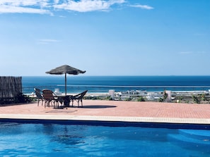 A poolside area with a view of the ocean, a table with chairs, and a thatched umbrella.