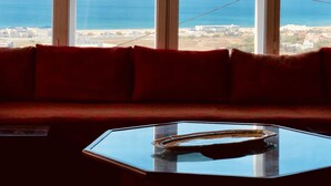 A room with a red sofa and a glass table, large windows with a view of the sea and buildings. - Villa Dar Jbila (Tangier)
