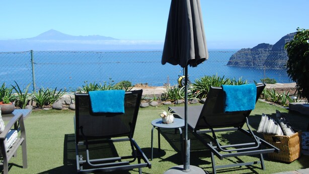Un patio con dos sillones de descanso, una mesa y una sombrilla, con vistas a la costa y una montaña al fondo.