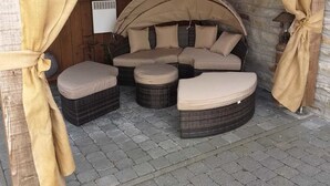 A covered outdoor seating area with wicker furniture and beige cushions, featuring a stone wall backdrop and a rustic wooden ceiling. - B&B La Petite Suisse (Malmedy)