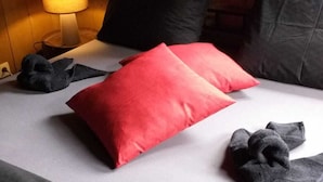 A bed with a red pillow, black pillows, and grey blankets. - B&B La Petite Suisse (Malmedy)