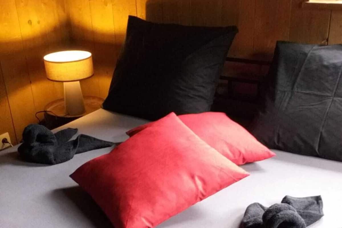 Un lit avec un oreiller rouge, des oreillers noirs et des couvertures grises.
